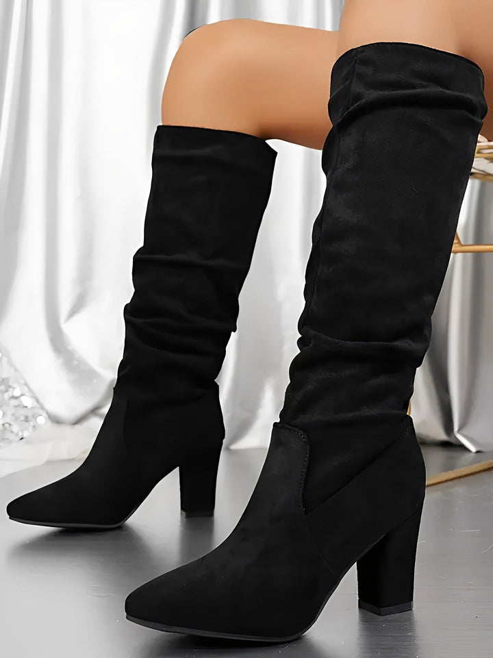 Bottes hautes en daim noir à talons épais, avec une tige plissée et une finition élégante, portées par une femme assise sur une chaise, photographiées en intérieur devant un rideau satiné.