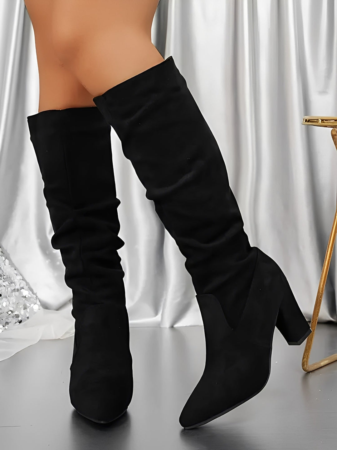 Bottes hautes en daim noir à talons épais, avec une tige légèrement plissée et une allure élégante, portées par une femme debout sur un sol brillant, devant un rideau satiné clair.