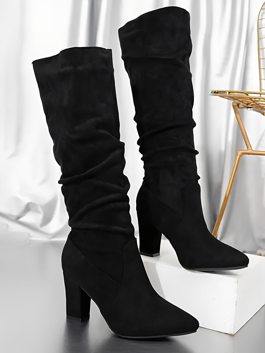 Paire de bottes hautes en daim noir à talons épais, avec une tige plissée et une silhouette élégante, présentées debout sur un socle blanc dans un décor intérieur aux rideaux satinés.
