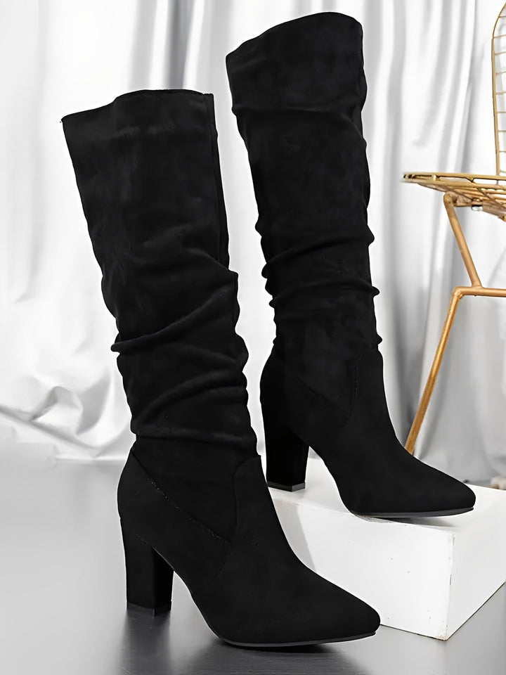 Paire de bottes hautes en daim noir à talons épais, avec une tige plissée et une silhouette élégante, présentées debout sur un socle blanc dans un décor intérieur aux rideaux satinés.