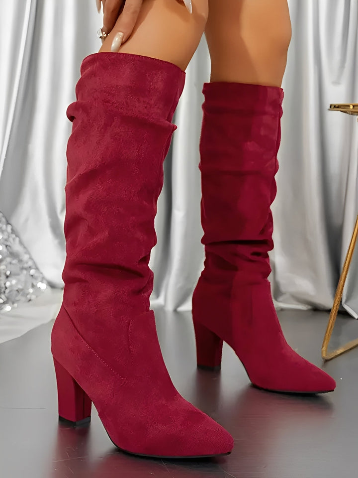 Bottes hautes en daim rouge vif à talons épais, avec une tige plissée et une finition élégante, portées par une femme debout sur un sol brillant, devant un rideau satiné argenté.