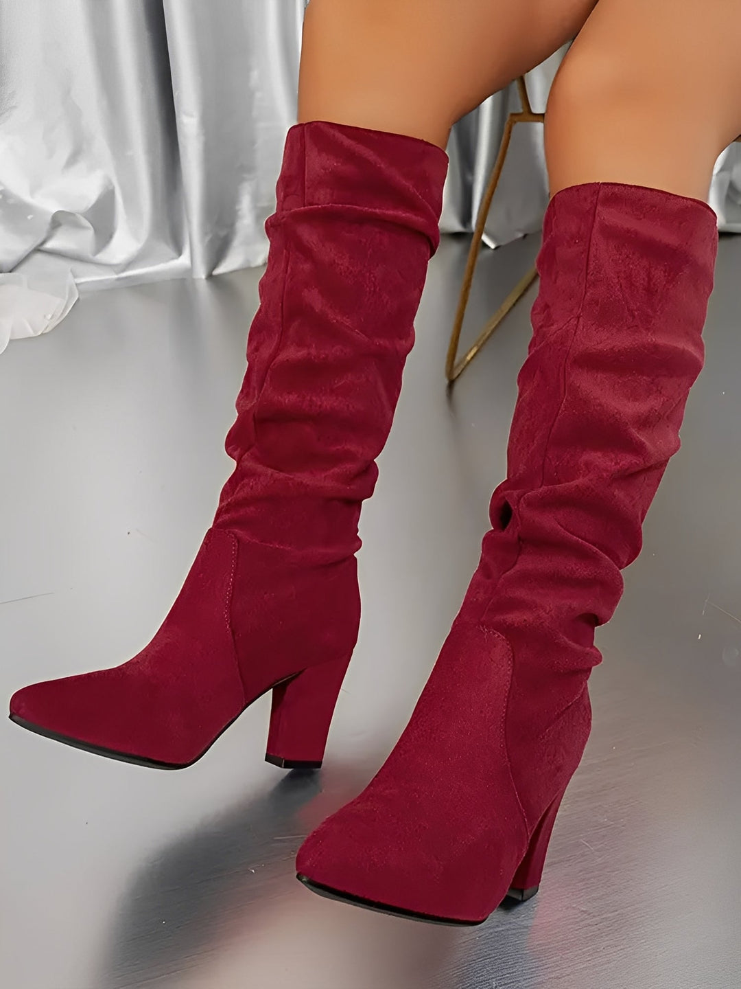 Bottes hautes en daim rouge à talons épais, avec une tige plissée et une coupe élégante, portées par une femme assise sur une chaise, photographiées en intérieur sur un sol brillant avec un rideau satiné en arrière-plan.