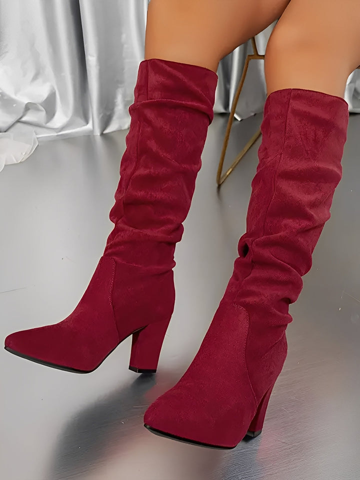 Bottes hautes en daim rouge à talons épais, avec une tige plissée et une coupe élégante, portées par une femme assise sur une chaise, photographiées en intérieur sur un sol brillant avec un rideau satiné en arrière-plan.