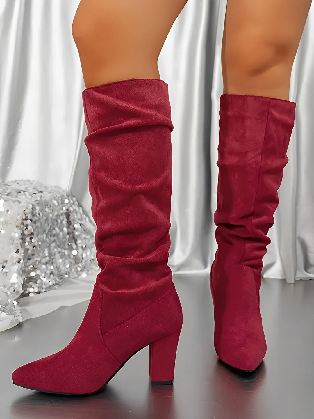 Bottes hautes en daim rouge à talons épais, avec une tige plissée et une allure élégante, portées par une femme debout sur un sol brillant, devant un rideau satiné argenté et une décoration scintillante.