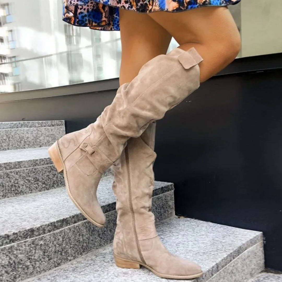 Bottes à talon femme Fabienne modernes en suédine beige, tige haute plissée jusqu’au genou avec nœud, fermeture zippée sur le côté et détail boucle, bout rond et petit talon, portées sur des marches.