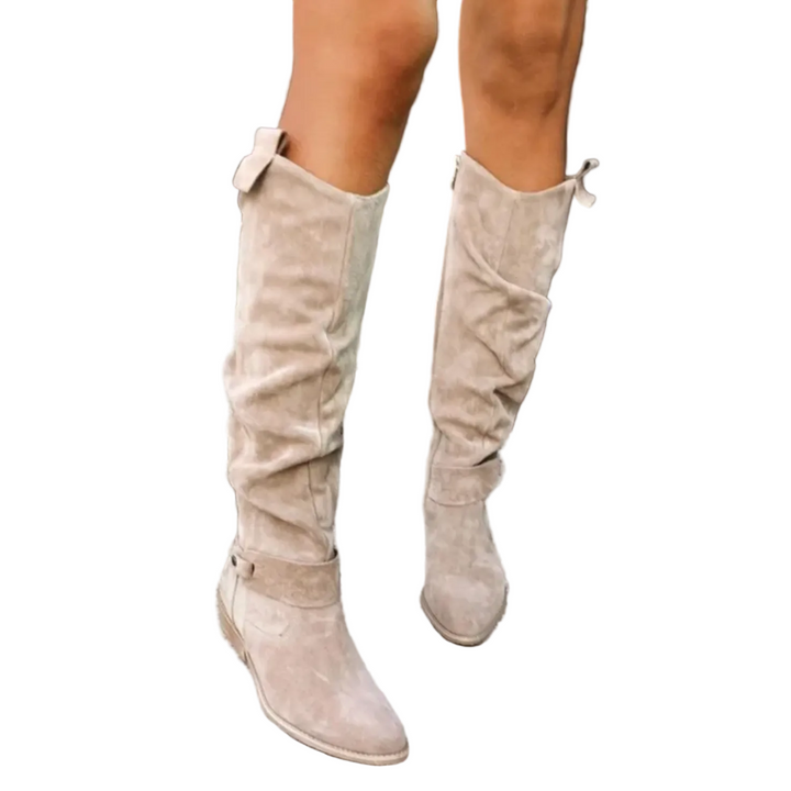Bottes à talon femme Fabienne modernes en suédine beige, tige haute plissée jusqu’au genou avec nœud à l’arrière, bout rond et petit talon, portées sur des marches en pierre.