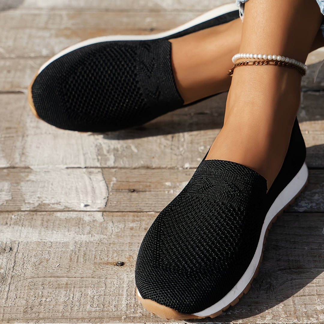 Chaussures d’été femme Fantine tendance noires portées aux pieds, slip-on en maille respirante, bout arrondi et semelle blanche légère avec dessous cranté, vue rapprochée sur terrasse en bois.