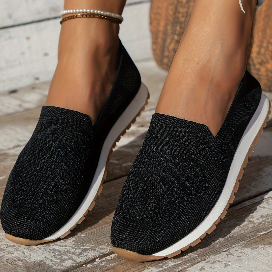 Chaussures d’été femme Fantine tendance noires, slip-on en maille respirante, bout arrondi et semelle blanche avec dessous cranté, portées en extérieur sur terrasse en bois.