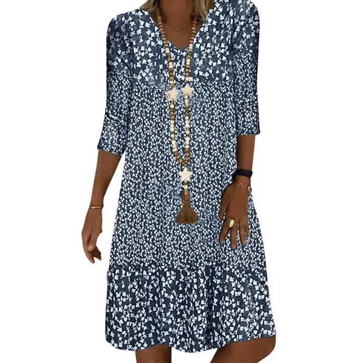 Robe midi femme Faustine moderne à imprimé floral bleu et blanc, manches 3/4 et col en V, coupe fluide avec bas volanté, portée avec un long collier, en extérieur.