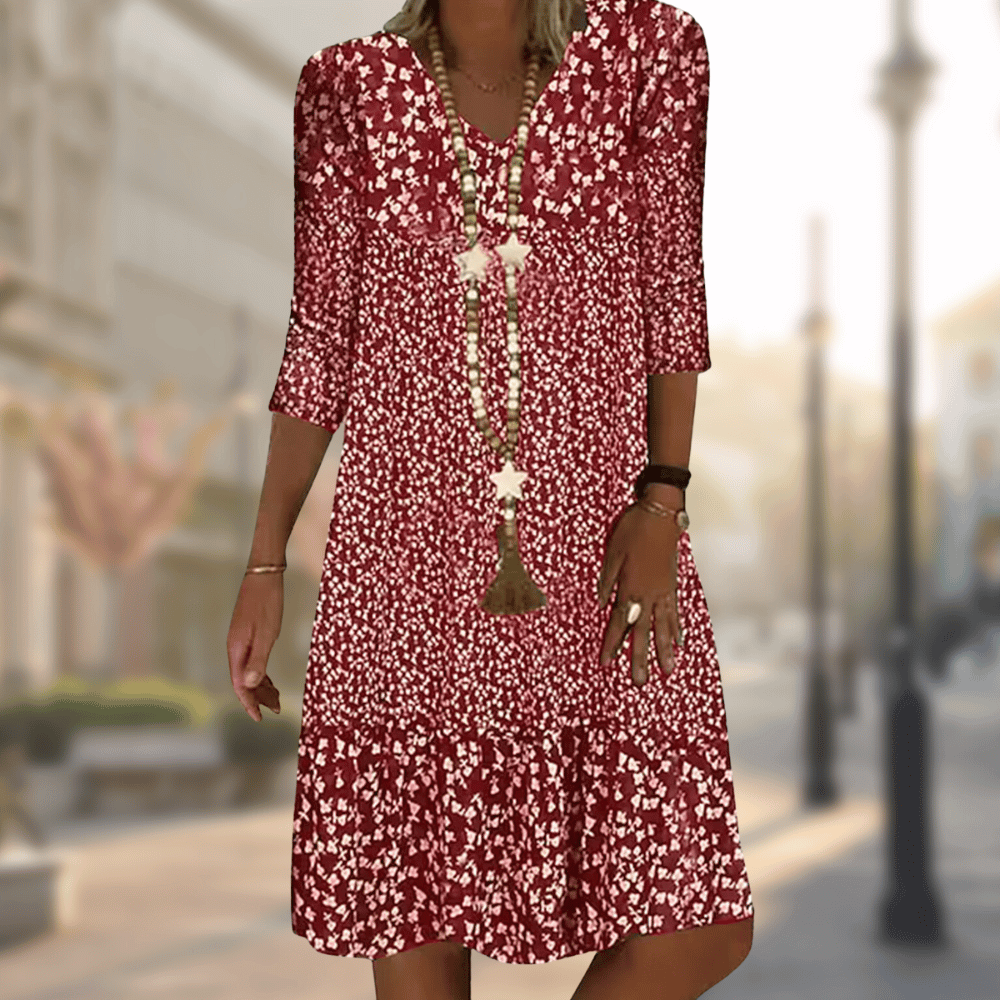 Robe midi femme Faustine moderne à imprimé floral rouge et blanc, manches 3/4 et col en V, coupe fluide avec bas volanté, portée avec un long collier, en extérieur.