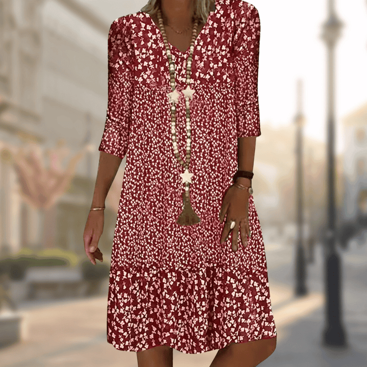 Robe midi femme Faustine moderne à imprimé floral rouge et blanc, manches 3/4 et col en V, coupe fluide avec bas volanté, portée avec un long collier, en extérieur.
