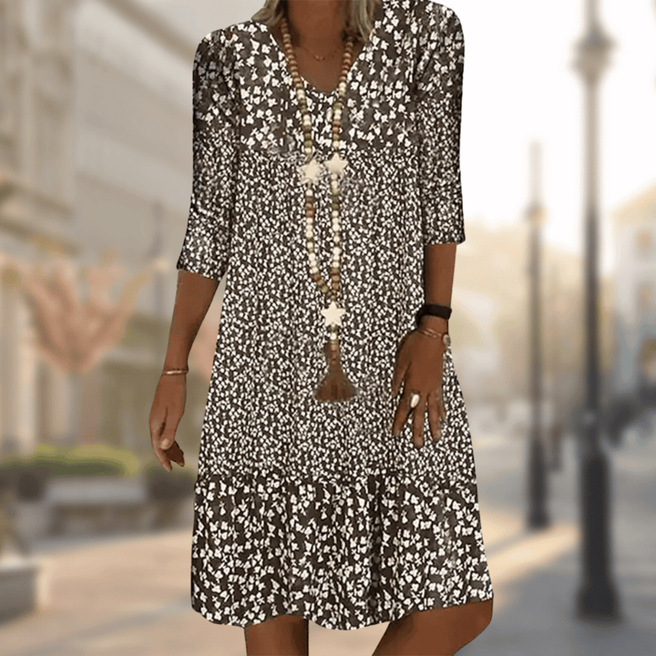 Robe midi femme Faustine moderne à imprimé floral noir et blanc, manches 3/4 et col en V, coupe fluide avec bas volanté, portée avec un long collier, en extérieur.