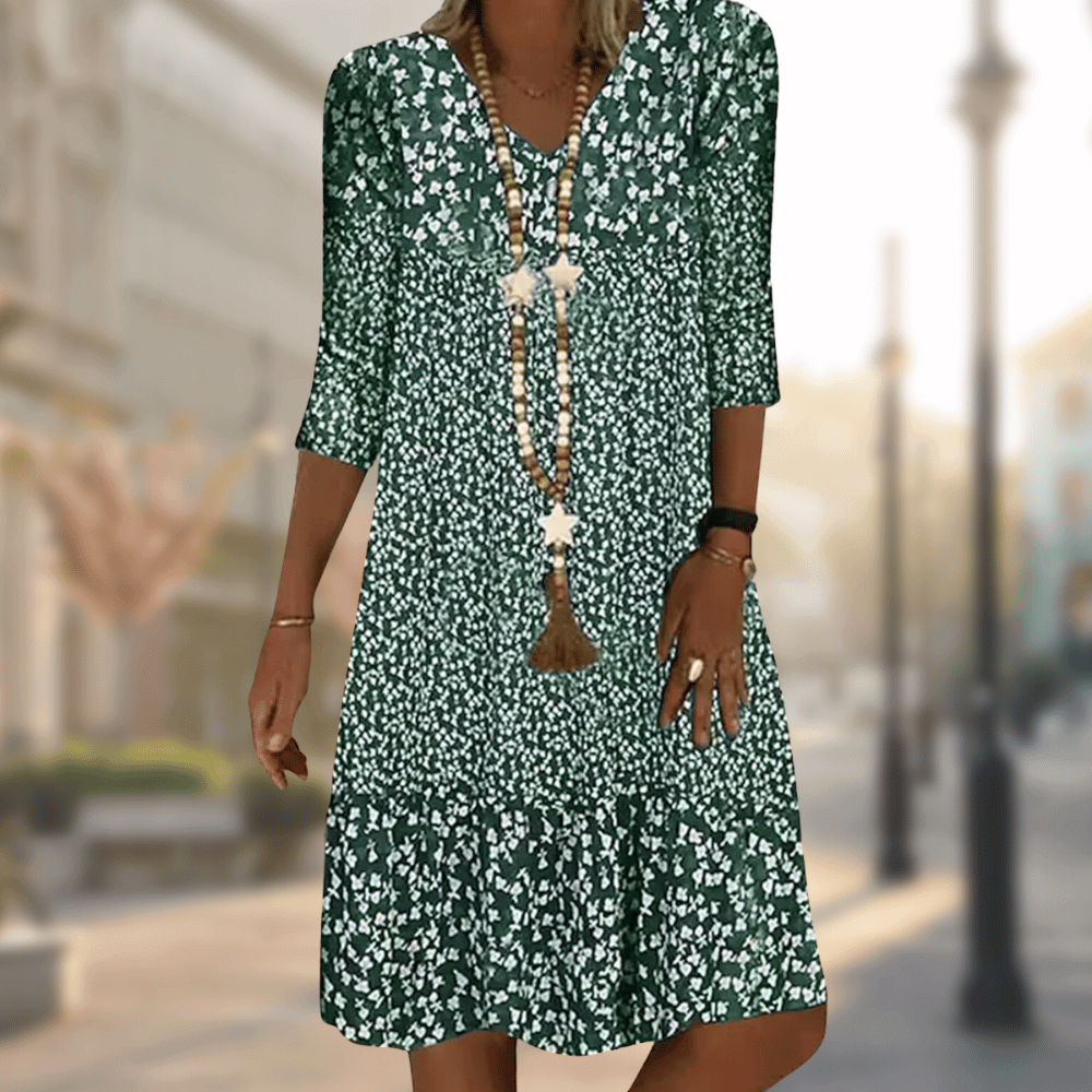 Robe midi femme Faustine moderne à imprimé floral vert et blanc, manches 3/4 et col en V, coupe fluide avec bas volanté, portée avec un long collier, en extérieur.