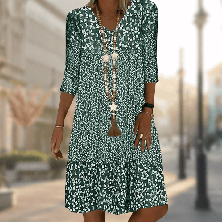 Robe midi femme Faustine moderne à imprimé floral vert et blanc, manches 3/4 et col en V, coupe fluide avec bas volanté, portée avec un long collier, en extérieur.