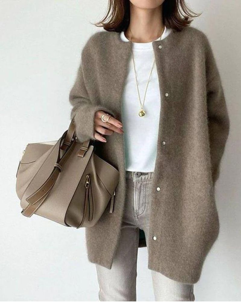 Femme portant un cardigan long couleur taupe à coupe droite, avec boutons sur le devant et texture douce et chaleureuse, porté sur un haut blanc et un jean clair, accompagné d’un sac à main beige et de bijoux discrets, dans un style chic et minimaliste.