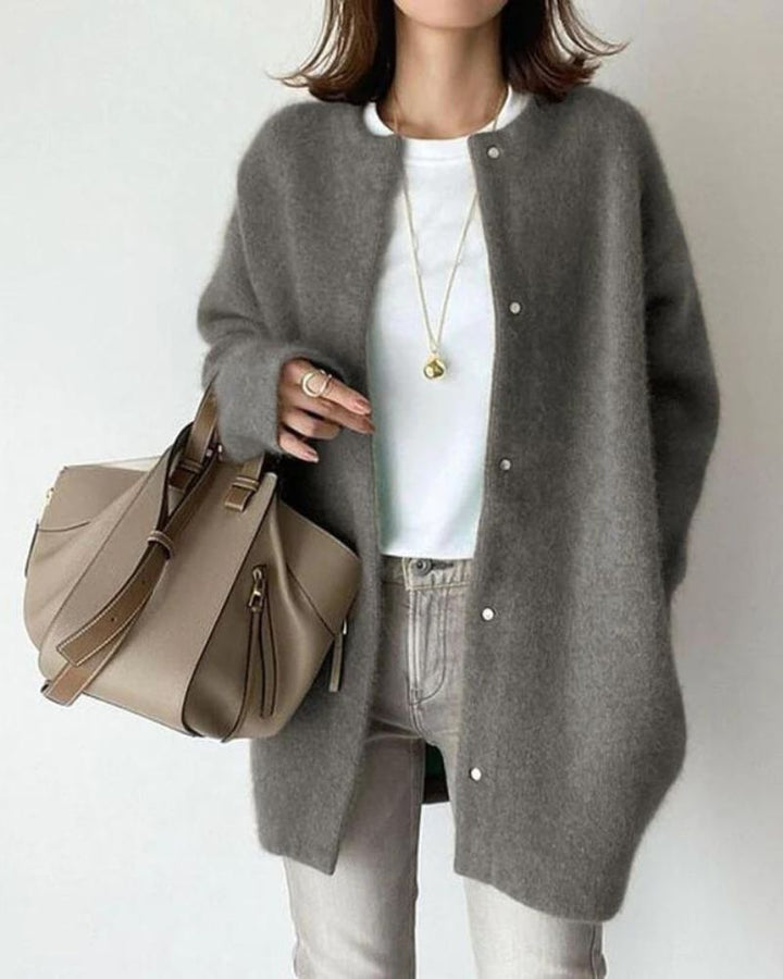 Femme portant un cardigan long gris à coupe droite, avec boutons sur le devant et texture douce et chaleureuse, porté sur un haut blanc et un jean clair, complété par un sac à main beige et des bijoux fins, dans un style chic et épuré.