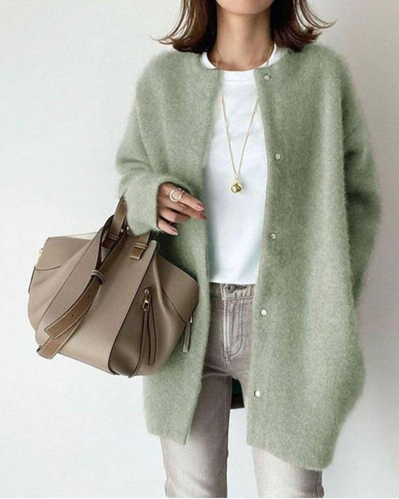 Femme portant un cardigan long vert sauge à coupe droite, avec boutons sur le devant et texture douce et chaude, porté sur un haut blanc et un jean clair, accompagné d’un sac à main beige et de bijoux délicats, dans un style élégant et naturel.