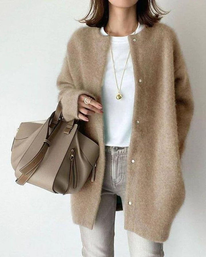 Femme portant un cardigan long beige chiné à coupe droite, avec boutons sur le devant et texture douce et chaude, porté sur un haut blanc et un jean clair, accessoirisé d’un sac à main beige et de bijoux délicats, dans un style chic et intemporel.