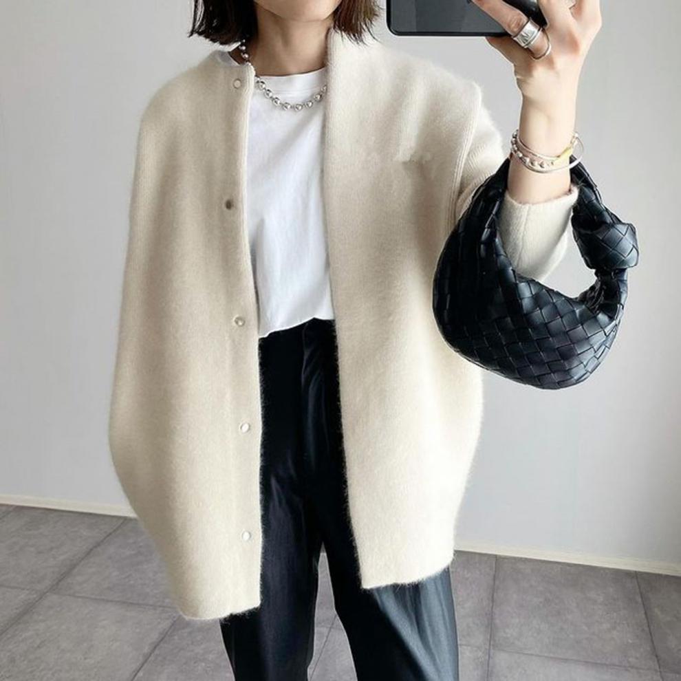 Femme portant un cardigan long écru à coupe droite, avec boutons sur le devant et texture douce et chaude, porté sur un t-shirt blanc et un pantalon noir, accessoirisé d’un sac à main noir tressé et de bijoux argentés, dans un style moderne et élégant.