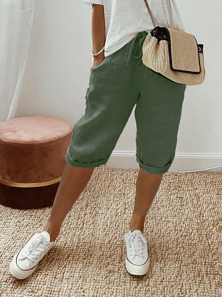 Pantalon pour femme de couleur vert olive avec longueur mi-mollet, coupe décontractée, taille élastique avec cordon de serrage, poches latérales, porté avec un t-shirt clair, des baskets blanches et un sac bandoulière en paille, vue de face des hanches jusqu’aux pieds dans un intérieur lumineux.