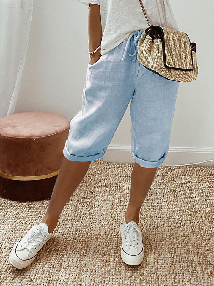 Pantalon pour femme de couleur bleu clair avec longueur mi-mollet, coupe décontractée, taille élastique avec cordon de serrage, poches latérales, ourlets légèrement retroussés, porté avec un t-shirt clair, des baskets blanches et un sac bandoulière en paille, vue de face des hanches jusqu’aux pieds dans un intérieur lumineux.