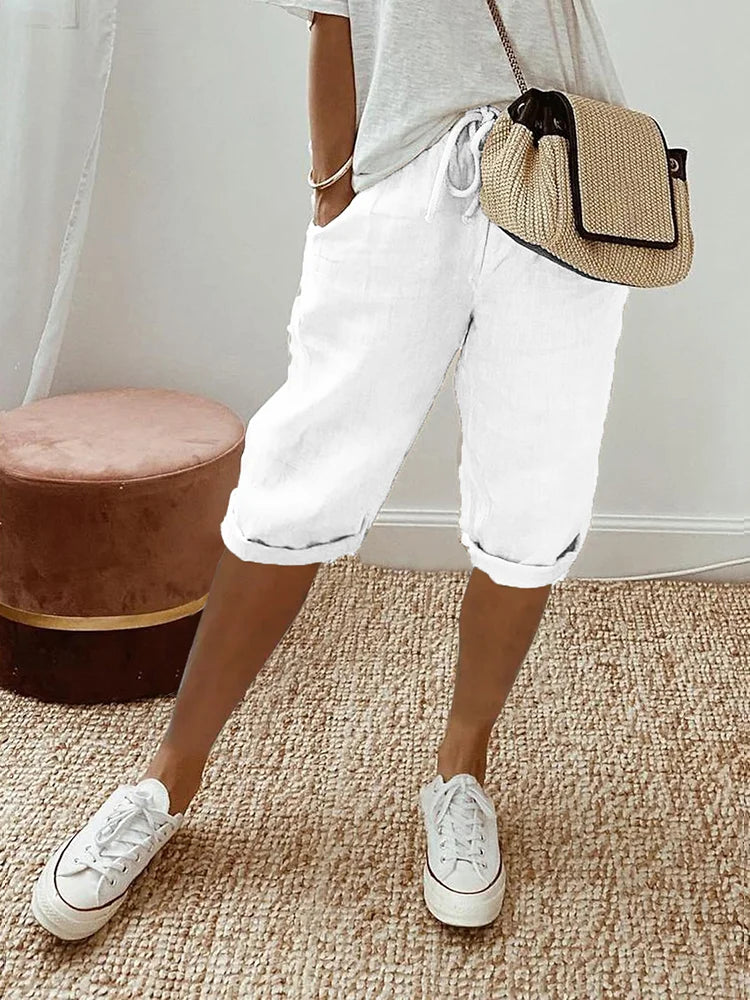 Pantalon pour femme de couleur blanche avec longueur mi-mollet, coupe décontractée, taille élastique avec cordon de serrage, poches latérales, ourlets légèrement retroussés, porté avec un t-shirt clair, des baskets blanches et un sac bandoulière en paille, vue de face des hanches jusqu’aux pieds dans un intérieur lumineux.