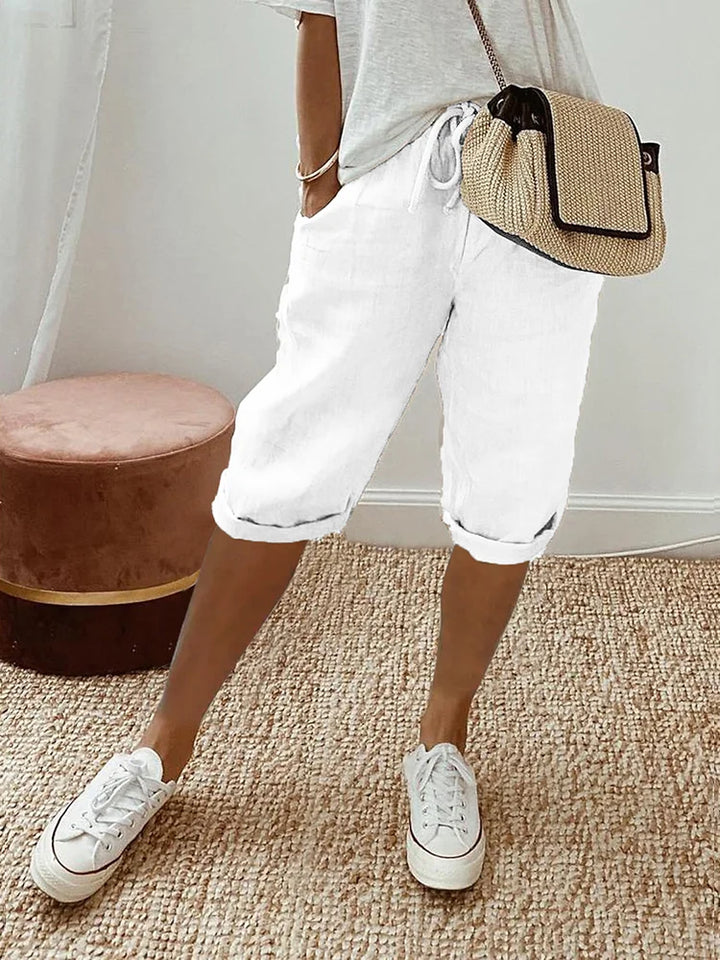 Pantalon pour femme de couleur blanche avec longueur mi-mollet, coupe décontractée, taille élastique avec cordon de serrage, poches latérales, ourlets légèrement retroussés, porté avec un t-shirt clair, des baskets blanches et un sac bandoulière en paille, vue de face des hanches jusqu’aux pieds dans un intérieur lumineux.