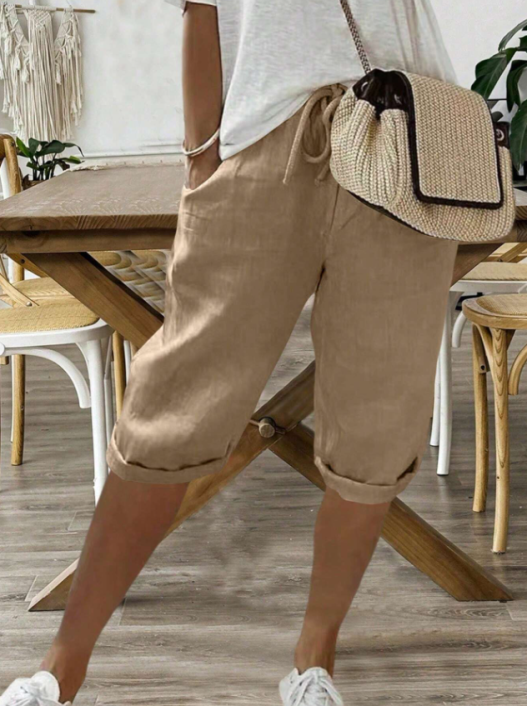 Pantalon pour femme de couleur beige naturel avec longueur mi-mollet, coupe décontractée et fluide, taille élastique avec cordon de serrage, poches latérales, ourlets retroussés, porté avec un t-shirt clair, des baskets blanches et un sac bandoulière en paille, vue de face des hanches jusqu’aux pieds dans un intérieur au style naturel.