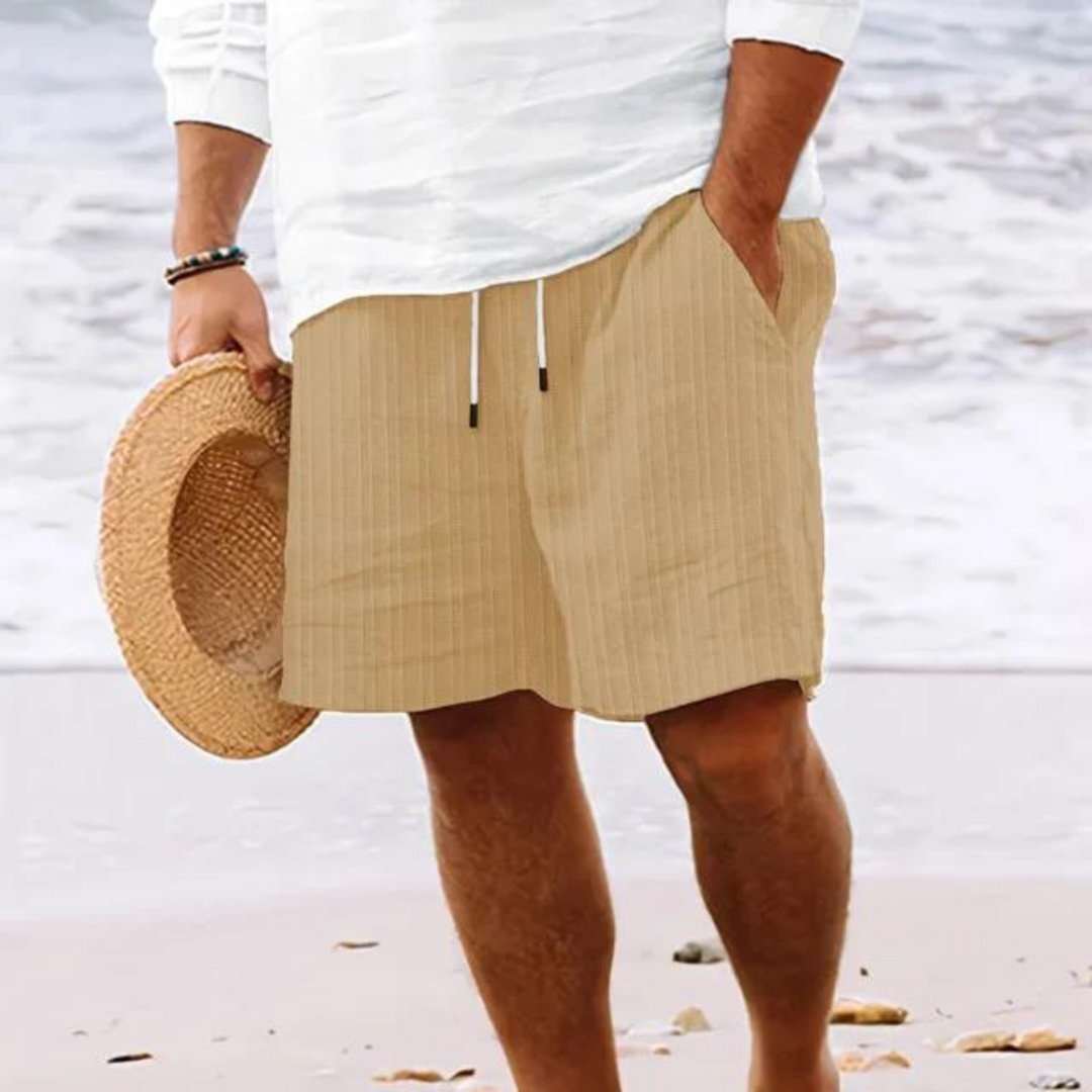 Un homme portant un short à rayures beiges avec poches et cordon de serrage, parfait pour un look décontracté et moderne à la plage.