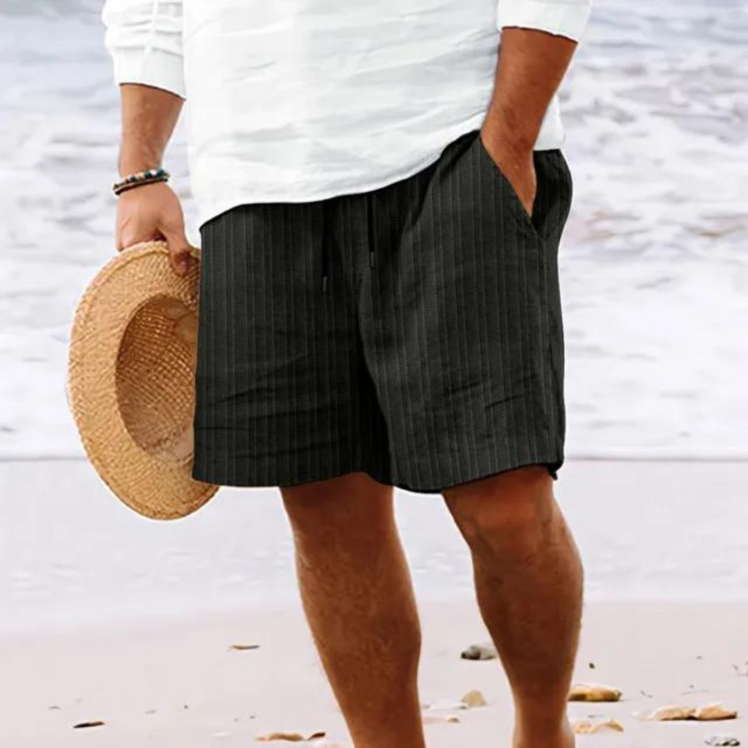Un homme portant un short à rayures noires avec poches et cordon de serrage, idéal pour un look décontracté et moderne à la plage.