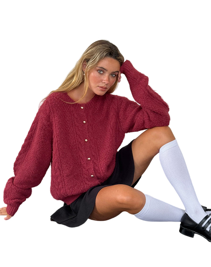 Femme assise portant un cardigan bordeaux en maille douce avec des boutons nacrés sur le devant. Le vêtement présente une coupe ample et un style confortable, idéal pour un look hivernal tendance. Elle l’associe à une jupe noire courte, des chaussettes hautes blanches et des mocassins noirs, créant une tenue chic et moderne.