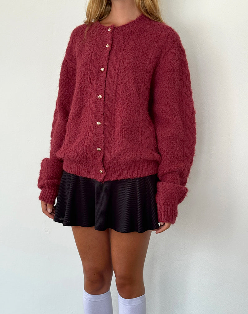 Femme portant un cardigan bordeaux en maille douce avec des boutons nacrés dorés sur le devant. Le vêtement présente une coupe ample et décontractée, offrant un style moderne et confortable. Elle l’associe à une jupe noire courte et des chaussettes hautes blanches, créant un look chic et tendance pour la saison automne-hiver.