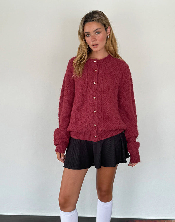 Femme debout portant un cardigan bordeaux en maille douce avec des boutons nacrés dorés sur le devant. Le vêtement présente une coupe légèrement ample, parfaite pour un style confortable et tendance. Elle l’associe à une jupe noire courte et des chaussettes hautes blanches, créant une tenue chic et moderne idéale pour la saison automne-hiver.