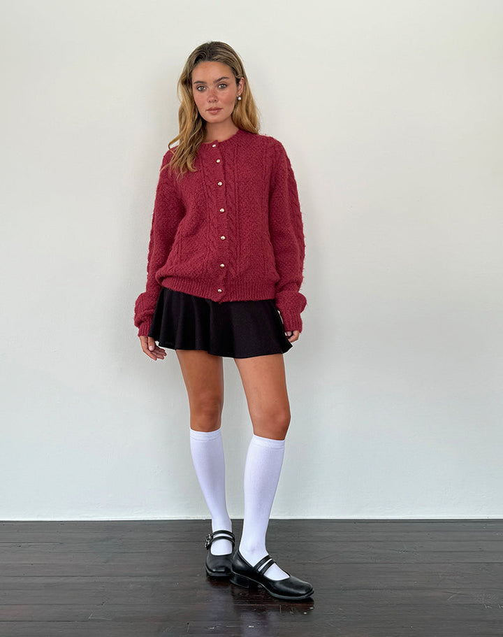 Femme debout portant un cardigan bordeaux en maille douce avec des boutons nacrés dorés sur le devant. Le vêtement présente une coupe ample et confortable, parfaite pour un look tendance et décontracté. Elle l’associe à une jupe noire courte, des chaussettes hautes blanches et des mocassins noirs, créant une tenue chic, moderne et élégante pour la saison automne-hiver.