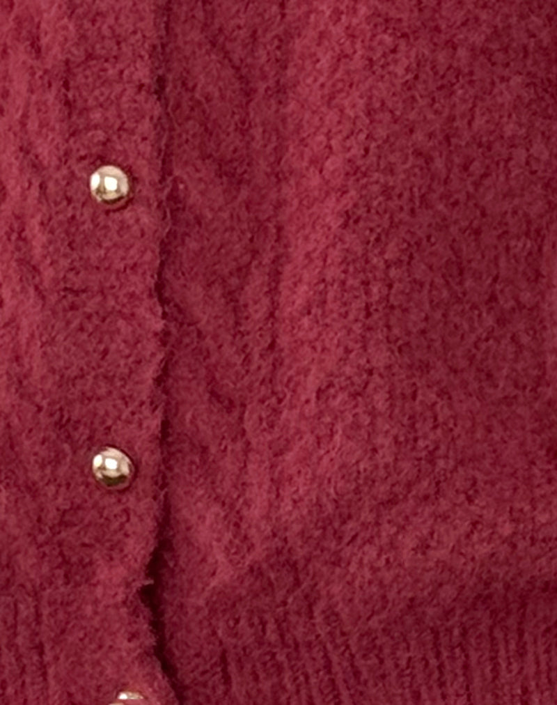 Gros plan sur la texture d’un cardigan bordeaux en maille épaisse. L’image met en valeur les détails du tricot doux et moelleux ainsi que les boutons nacrés dorés alignés sur le devant. Le tissu présente une finition torsadée subtile, soulignant la qualité et le style chaleureux du vêtement.