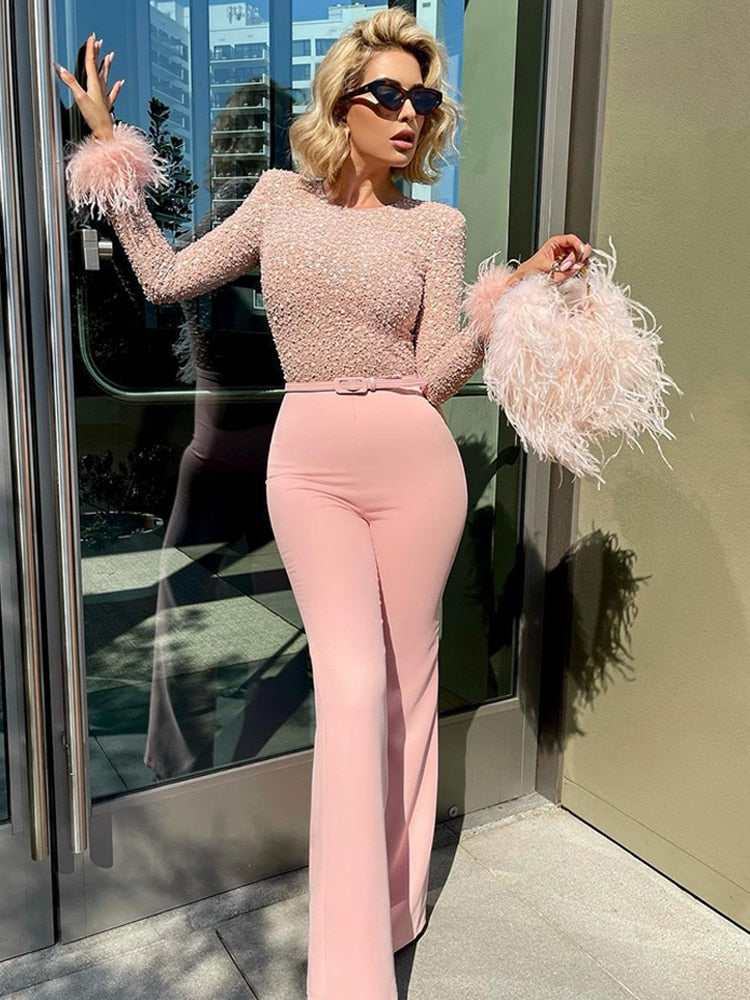 Une femme sophistiquée porte un haut transparent orné de sequins brillants, accompagné de plumes délicates sur les manches, créant un look glamour. Elle porte un pantalon taille haute rose pâle qui complète parfaitement l'ensemble, avec une silhouette élégante. L'arrière-plan moderne et lumineux accentue l'aspect chic de la tenue.