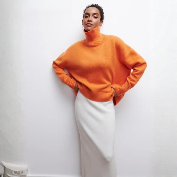 Pull oversize à col roulé pour femme, en orange vif, avec une coupe ample et des manches longues. Le col roulé est large et côtelé, tandis que le bas du pull présente également une finition côtelée. Ce pull est porté avec une jupe blanche, créant un look moderne et audacieux, parfait pour l'hiver.