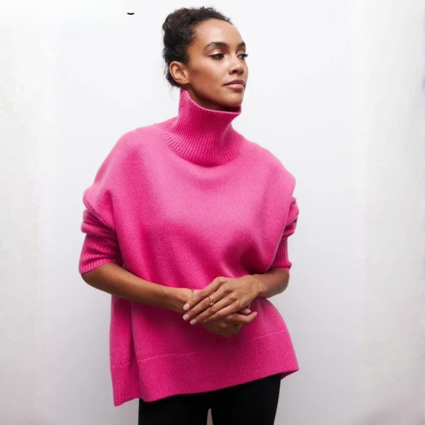 Pull oversize à col roulé pour femme, en rose vif, avec une coupe ample et des manches courtes. Le col roulé est large et côtelé, et le bas du pull présente également une finition côtelée. Ce pull est porté avec un pantalon noir, créant un look moderne et audacieux, parfait pour l'hiver.