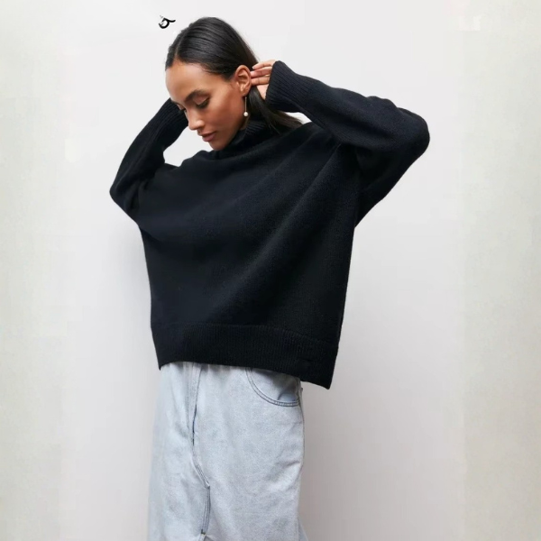 Pull oversize à col roulé pour femme, en noir, avec une coupe ample et des manches longues. Le col roulé est large et côtelé, et le bas du pull présente également une finition côtelée. Ce pull est porté avec une jupe en denim claire, créant un look élégant et décontracté, parfait pour l'hiver.