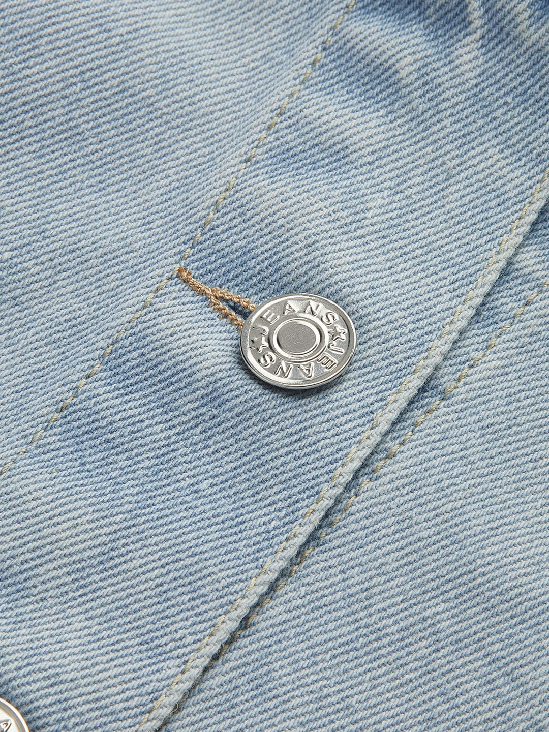Zoom sur le bouton métallique de la veste en jean courte Giselle. Ce bouton argenté avec l'inscription 'JEANS' ajoute un détail élégant et moderne à la veste, accentuant son style décontracté. Le denim bleu clair et la finition soignée de cette veste en font un choix idéal pour un look urbain tendance.