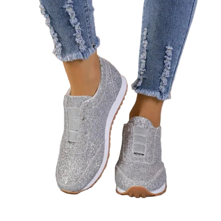 Baskets à paillettes pour femme Gwendolyn, coloris argenté scintillant, modèle slip-on avec bande élastique sur le dessus et semelle blanche avec gomme, portées avec un jean sur fond studio.