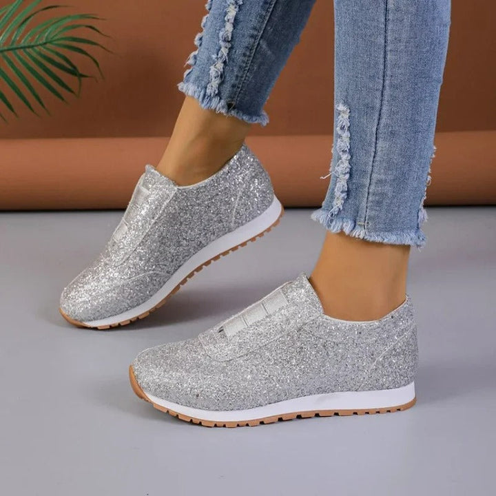 Baskets à paillettes pour femme Gwendolyn argentées, slip-on scintillantes avec bande élastique sur le dessus, semelle blanche et gomme crantée, vue de profil portées avec un jean sur fond studio.
