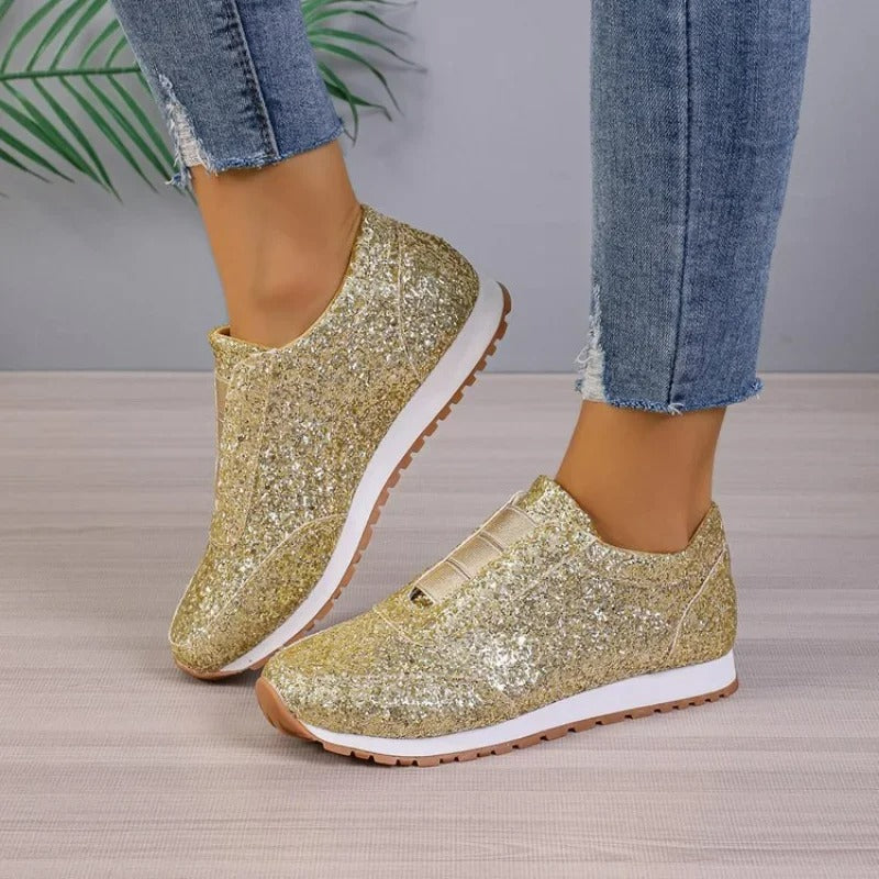 Baskets à paillettes pour femme Gwendolyn dorées, slip-on scintillantes avec bande élastique sur le dessus, semelle blanche et gomme crantée, vue en angle portées avec un jean sur fond studio.