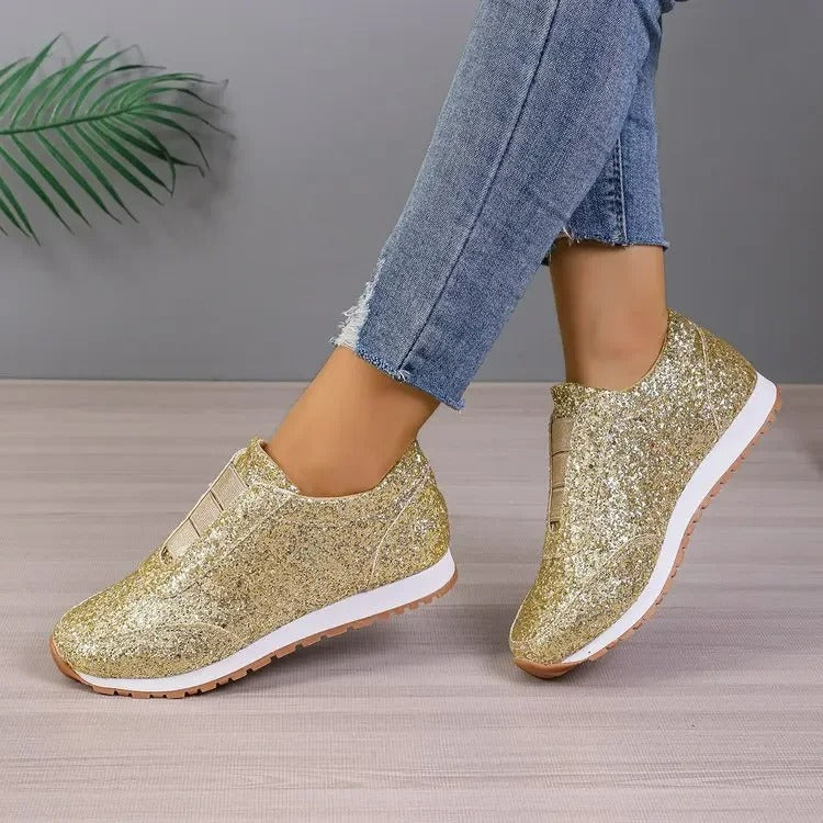 Baskets à paillettes pour femme Gwendolyn dorées, slip-on scintillantes avec bande élastique, semelle blanche et gomme crantée, portées avec un jean sur fond studio gris.