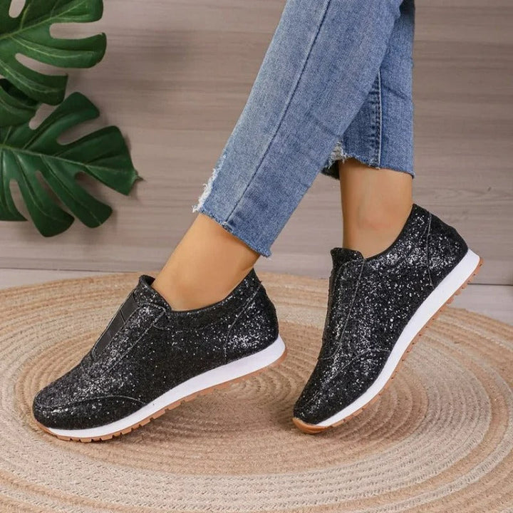 Baskets à paillettes pour femme Gwendolyn noires scintillantes, slip-on avec bande élastique, semelle blanche et gomme, portées avec un jean sur tapis rond en jute.