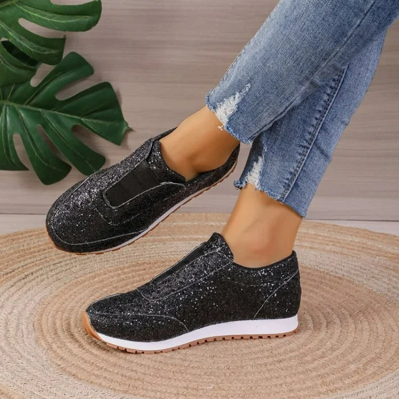 Baskets à paillettes pour femme Gwendolyn noires, slip-on scintillantes avec bande élastique, semelle blanche et gomme crantée, vue de profil portées avec un jean sur tapis en jute.