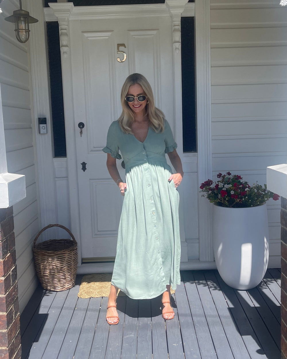 Robe midi d'été pour femme, portée par une modèle devant une porte. Style tendance avec des boutons sur le devant, des manches à volants et une coupe fluide, parfaite pour une journée estivale décontractée.