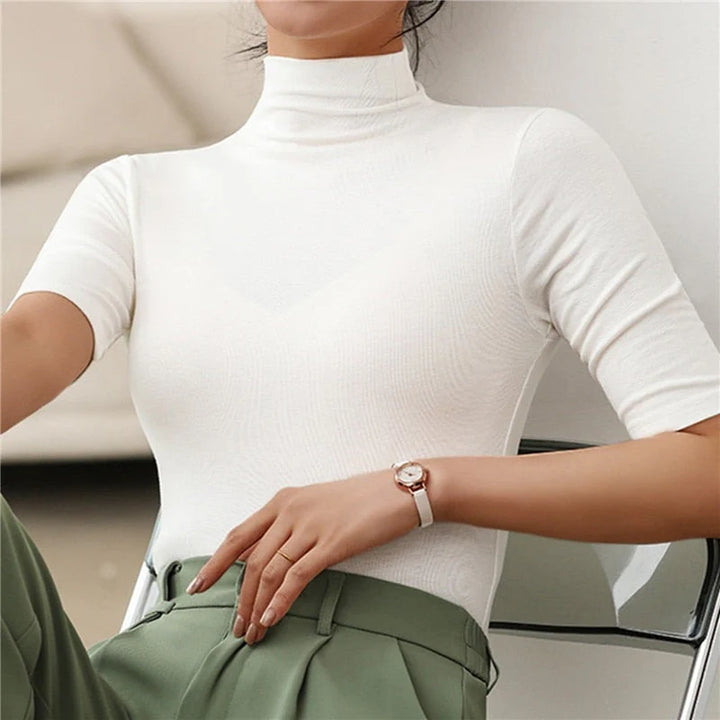 Femme portant un haut blanc à col roulé et manches courtes. Le vêtement est simple et élégant, offrant un look raffiné avec un ajustement près du corps. Elle associe le haut à un pantalon vert, créant une silhouette moderne et stylée, avec une montre élégante au poignet. Ce look est parfait pour un style décontracté ou une tenue de bureau.