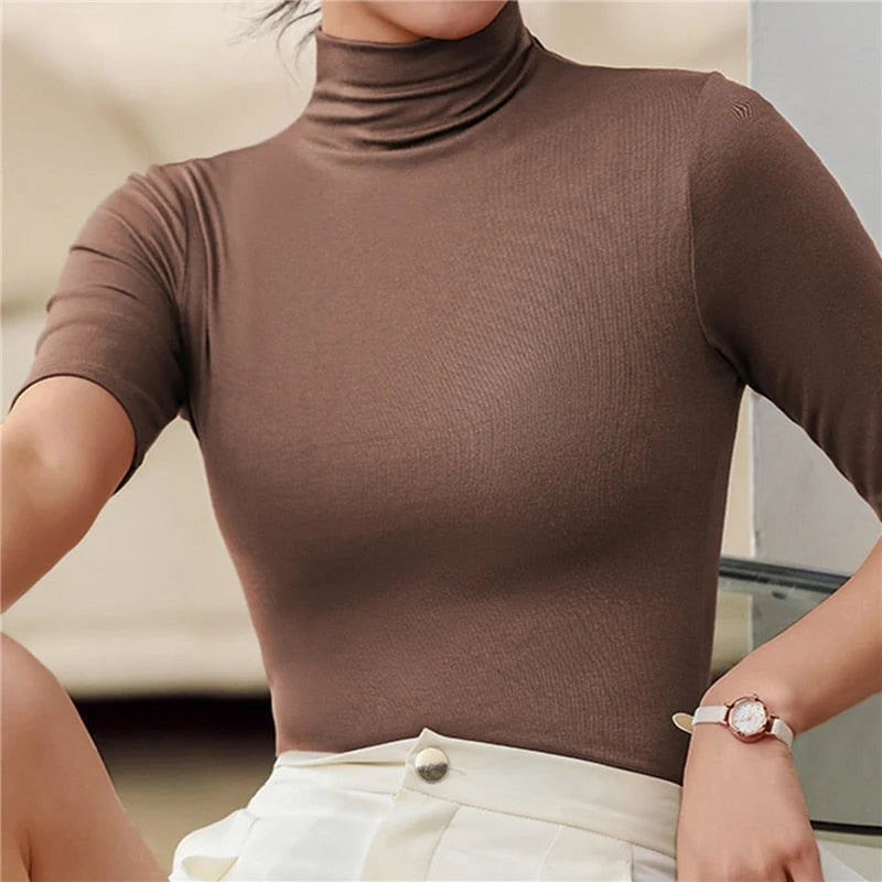Femme portant un haut moulant à col roulé et manches courtes de couleur marron. Le vêtement met en valeur sa silhouette avec son ajustement près du corps. Elle associe ce top avec une jupe blanche, complétée par une montre élégante au poignet, créant un look chic et moderne.