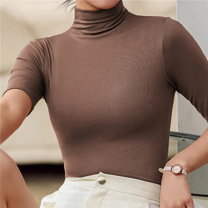 Femme portant un haut moulant à col roulé et manches courtes de couleur marron. Le vêtement met en valeur sa silhouette avec son ajustement près du corps. Elle associe ce top avec une jupe blanche, complétée par une montre élégante au poignet, créant un look chic et moderne.